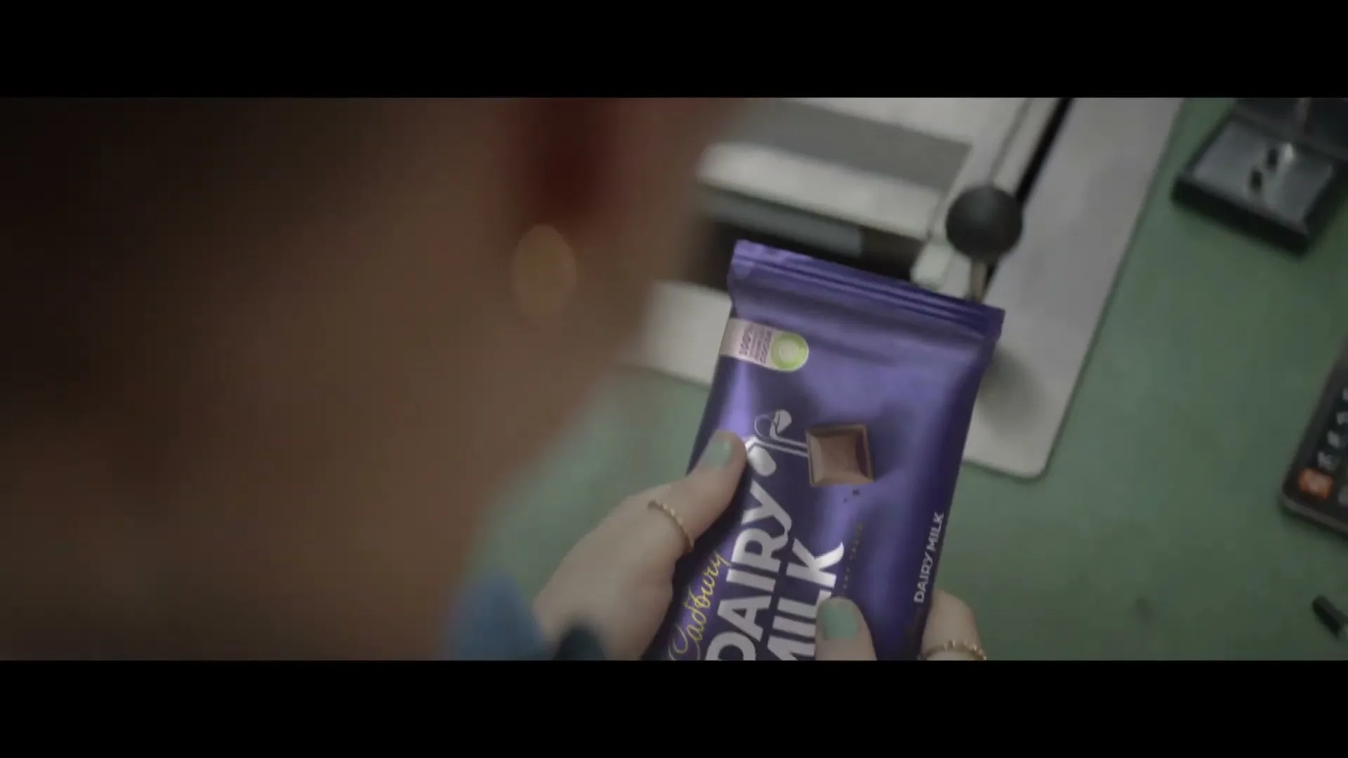 A person's hands hold a purple Cadbury Dairy Milk chocolate bar, with a calculator and other items visible on a green counter in a garage.