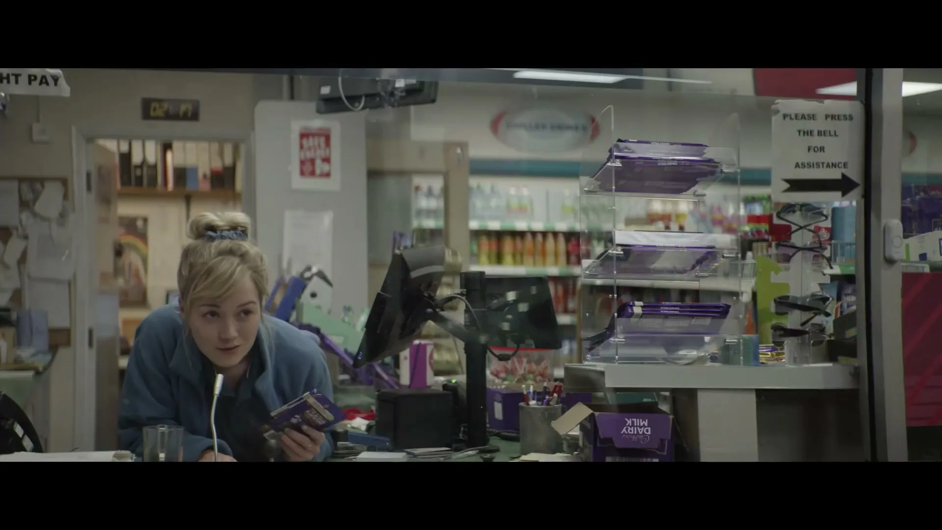 A young female cashier with blonde hair tied in a bun, wearing a blue fleece, looks up from scanning a Cadbury chocolate bar at a gas station convenience store.