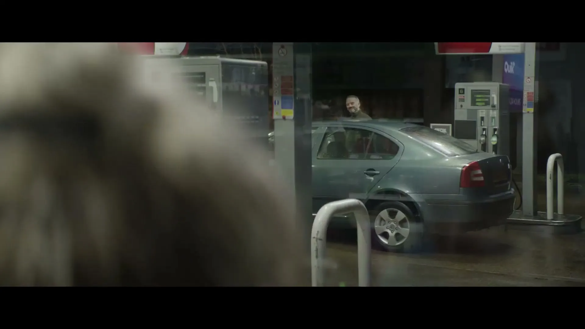 A man with a beard and a green jacket smiles while standing next to a car at a gas station at night, seen through a blurry foreground.
