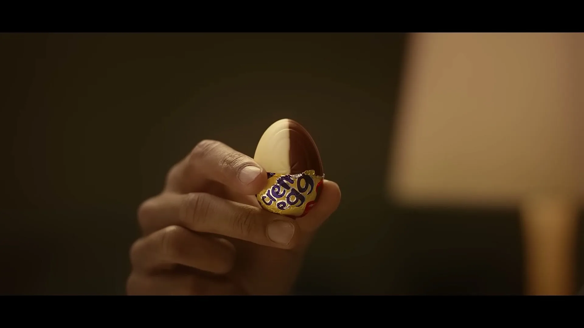 A person's hand holds a half-eaten Cadbury Creme Egg, revealing the white and yellow fondant inside, in a dimly lit room.