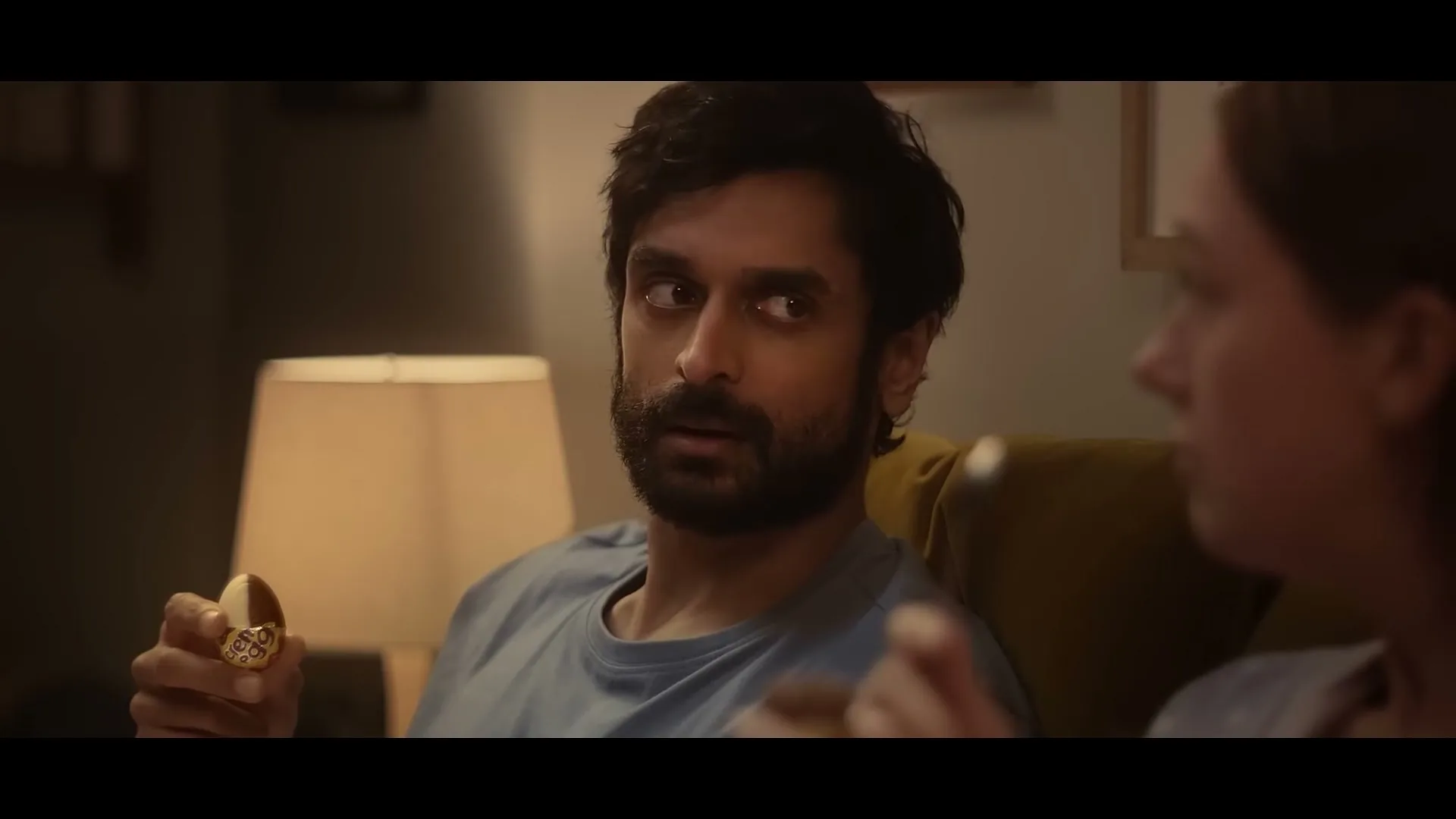 A man with a beard in a blue t-shirt holds a golden egg while talking to a woman on a couch in a dimly lit living room.