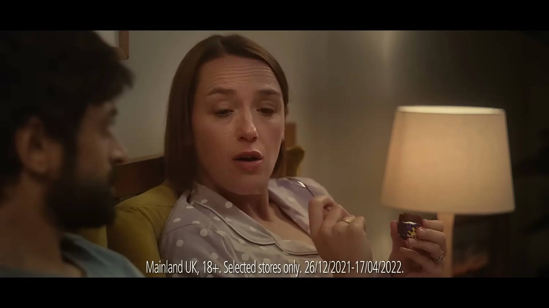 A woman in pajamas holds a chocolate egg and a fork, looking at a man next to her in a bedroom at night.