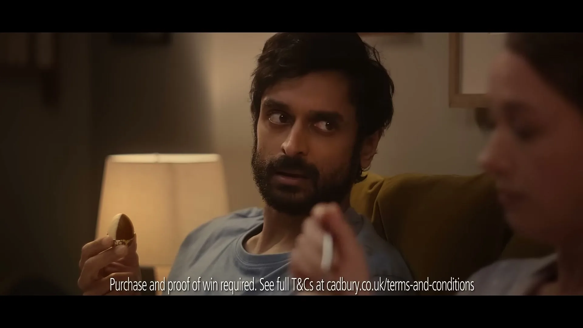 A man with a beard and a woman are sitting on a couch, eating chocolate, and talking in a dimly lit living room.