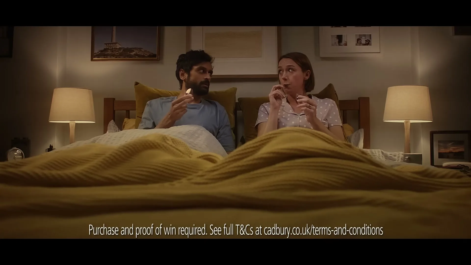 A man and a woman, both in their 30s, sit up in bed, each holding a Cadbury Creme Egg, looking at each other in a bedroom at night.