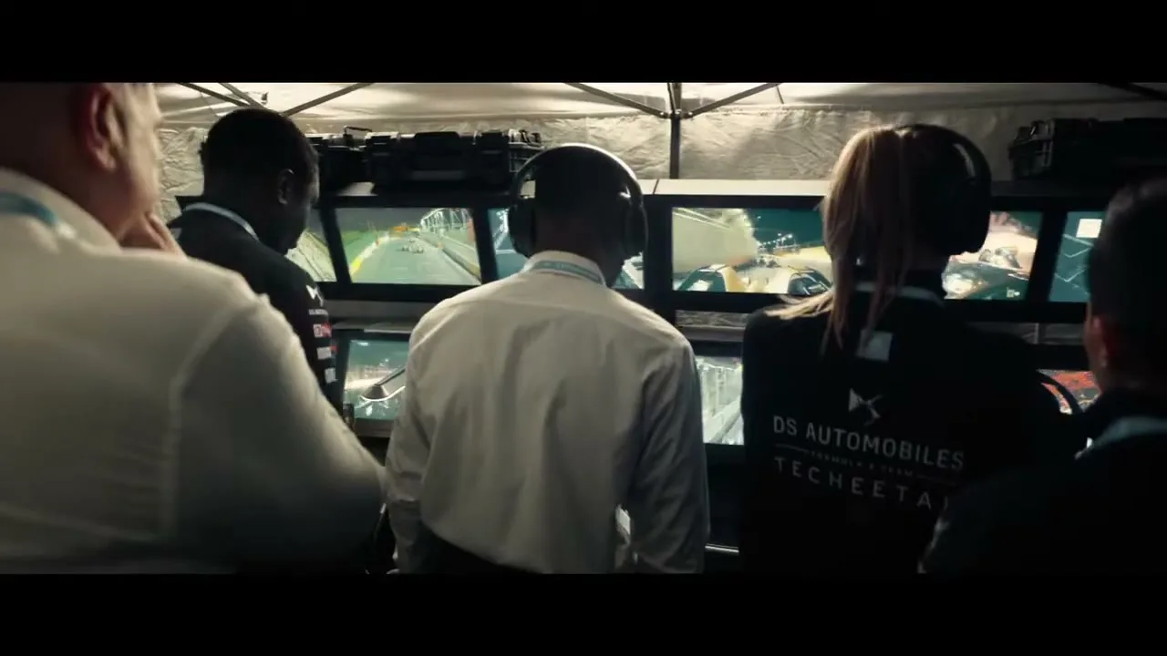 A group of diverse race team members, wearing headsets and professional attire, watch multiple screens displaying a Formula E race from inside a tent at night.