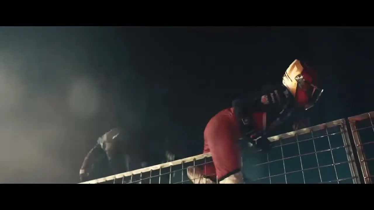 Two race car drivers, one in red and one in black, climb over a metal fence at night, with smoke in the background.