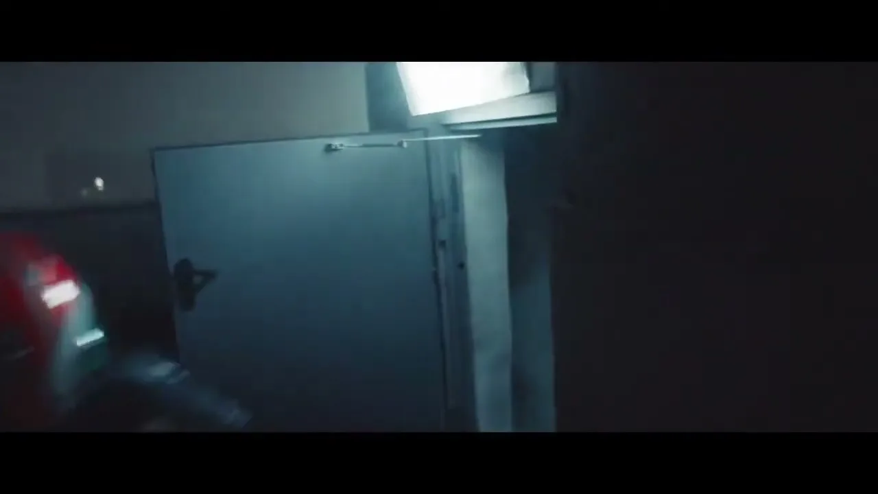 A person in a racing suit and helmet is seen from behind, running through a dark, industrial-looking hallway with a bright light source ahead.