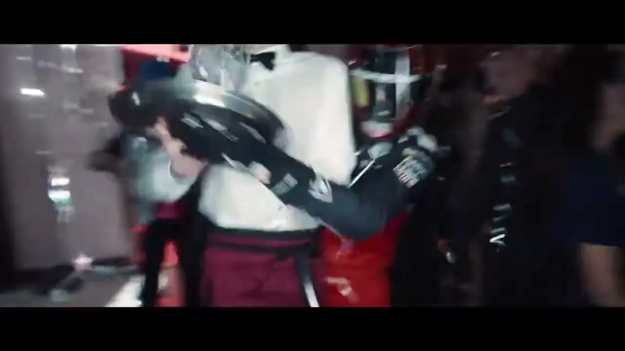 A Formula E race car driver in full gear, including a helmet, walks through a crowded, dimly lit party, surrounded by people dancing and holding drinks.