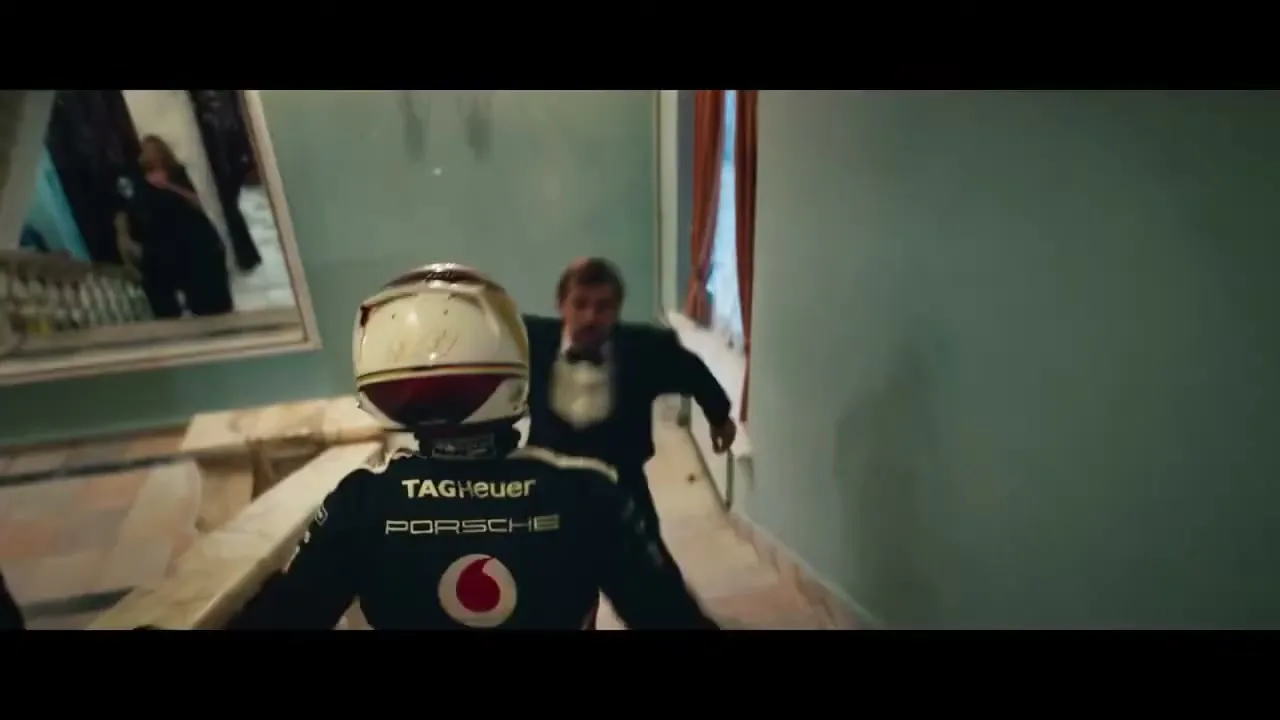 A race car driver in full gear, including a helmet, runs through a grand hall, pursued by a man in a tuxedo.
