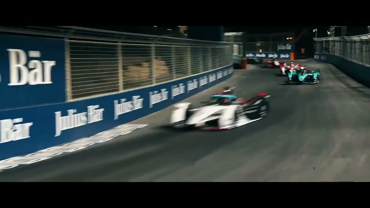 A white and black Formula E race car speeds around a track at night, with other cars visible in the background.