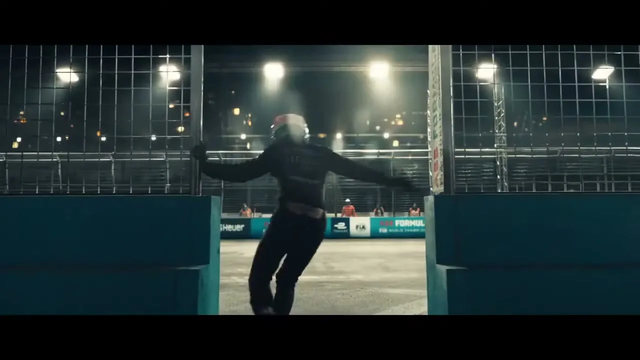 A race car driver in a black suit and helmet runs onto a brightly lit race track at night, seen through a gap in a fence.