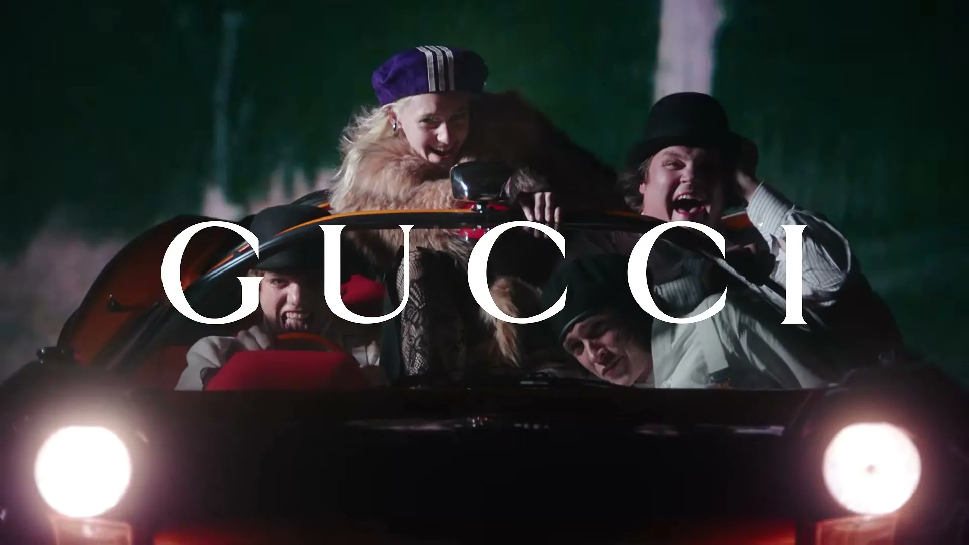 Four people, dressed in vintage clothing, are crammed into a small car, driving at night with the Gucci logo superimposed.