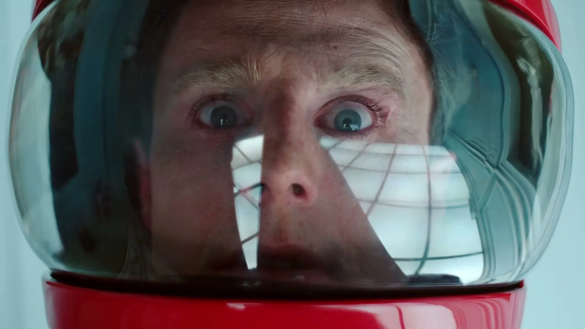 A man with blonde hair and blue eyes, wearing a red astronaut helmet, looks directly at the camera with a surprised expression in a bright, modern interior.