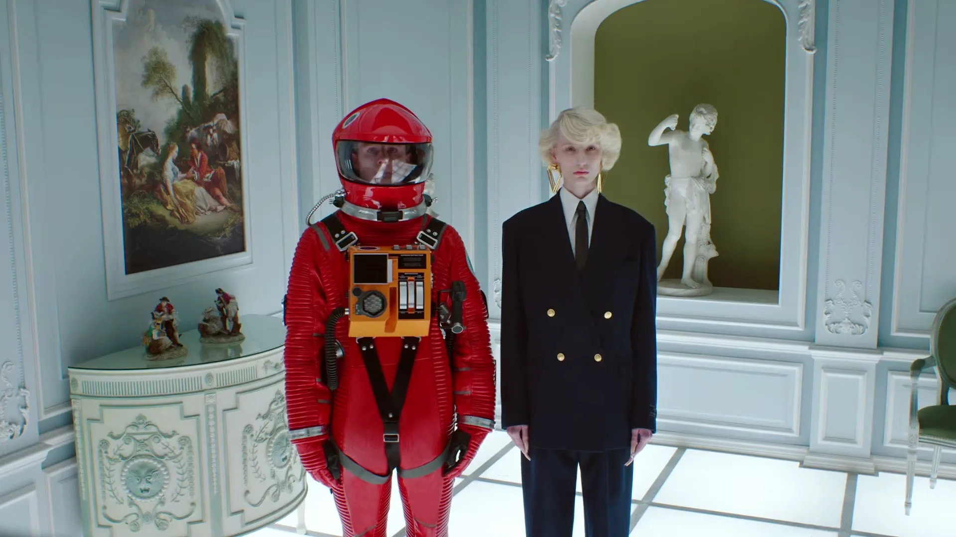 A person in a red astronaut suit stands next to a person in a dark suit with a blonde wig, both looking forward in a white, ornate room.