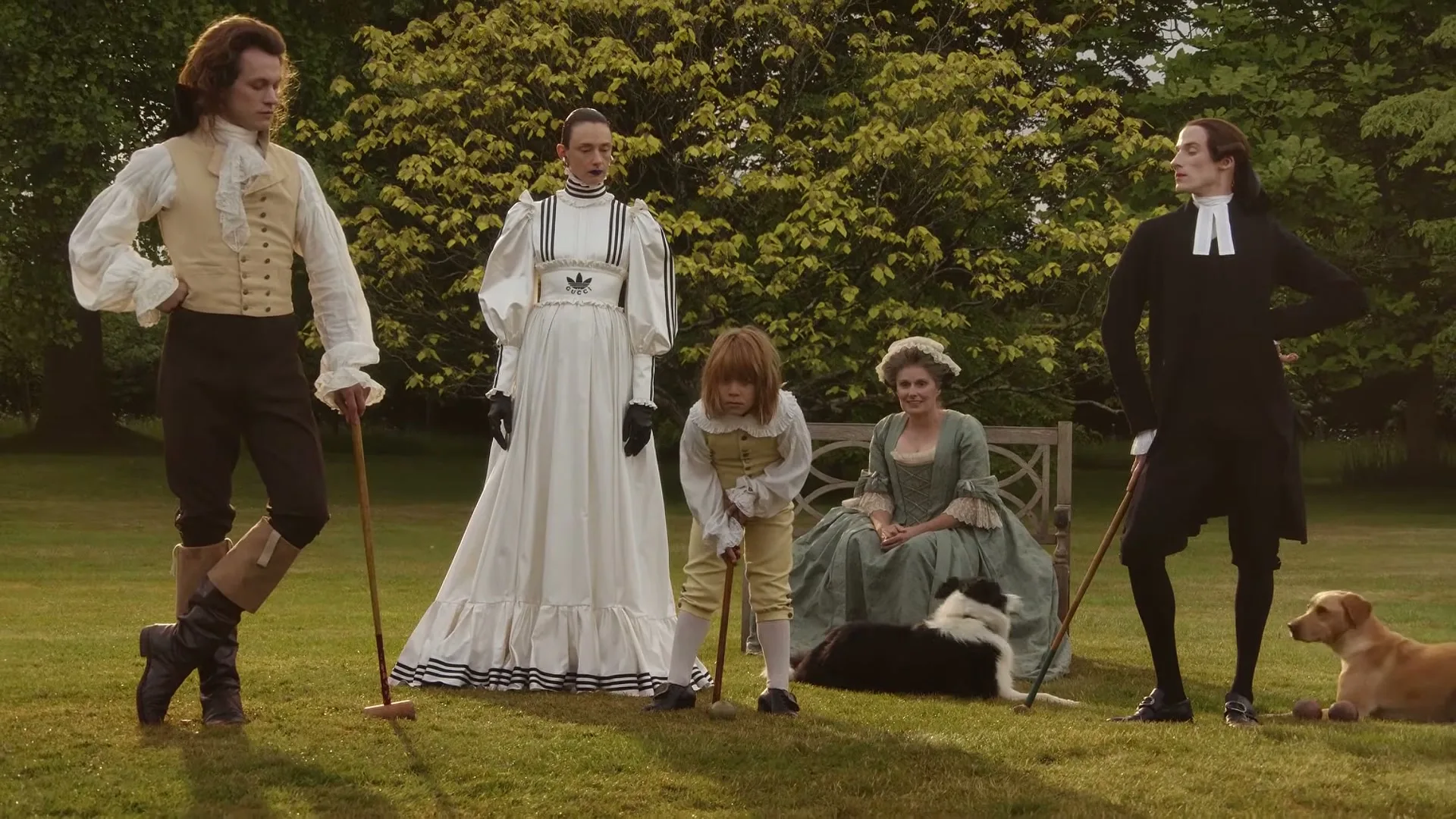 A group of people in period costumes, including a woman in a white dress with Adidas stripes, play croquet on a sunny lawn with two dogs.