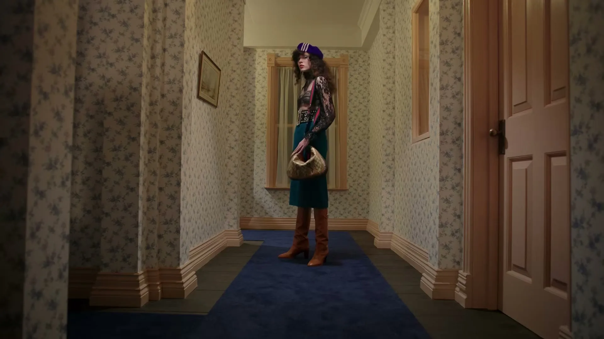 A young woman in a purple beret, patterned top, green skirt, and brown boots stands in a hallway with floral wallpaper, holding a small handbag.