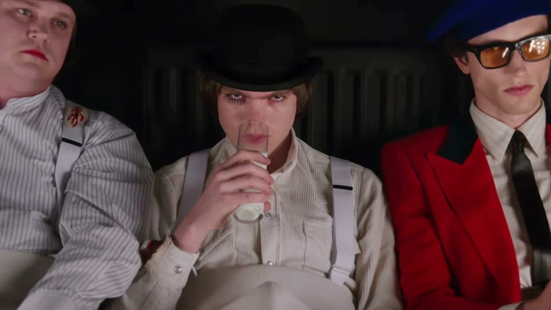 Three young men in stylized costumes sit in a dark interior, with the central figure drinking milk from a glass.