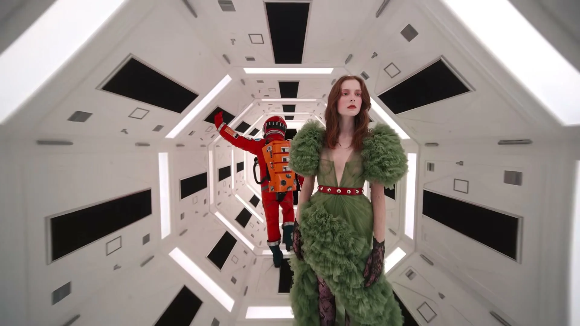 A woman in a green ruffled gown and a person in a red astronaut suit walk down a futuristic white corridor.