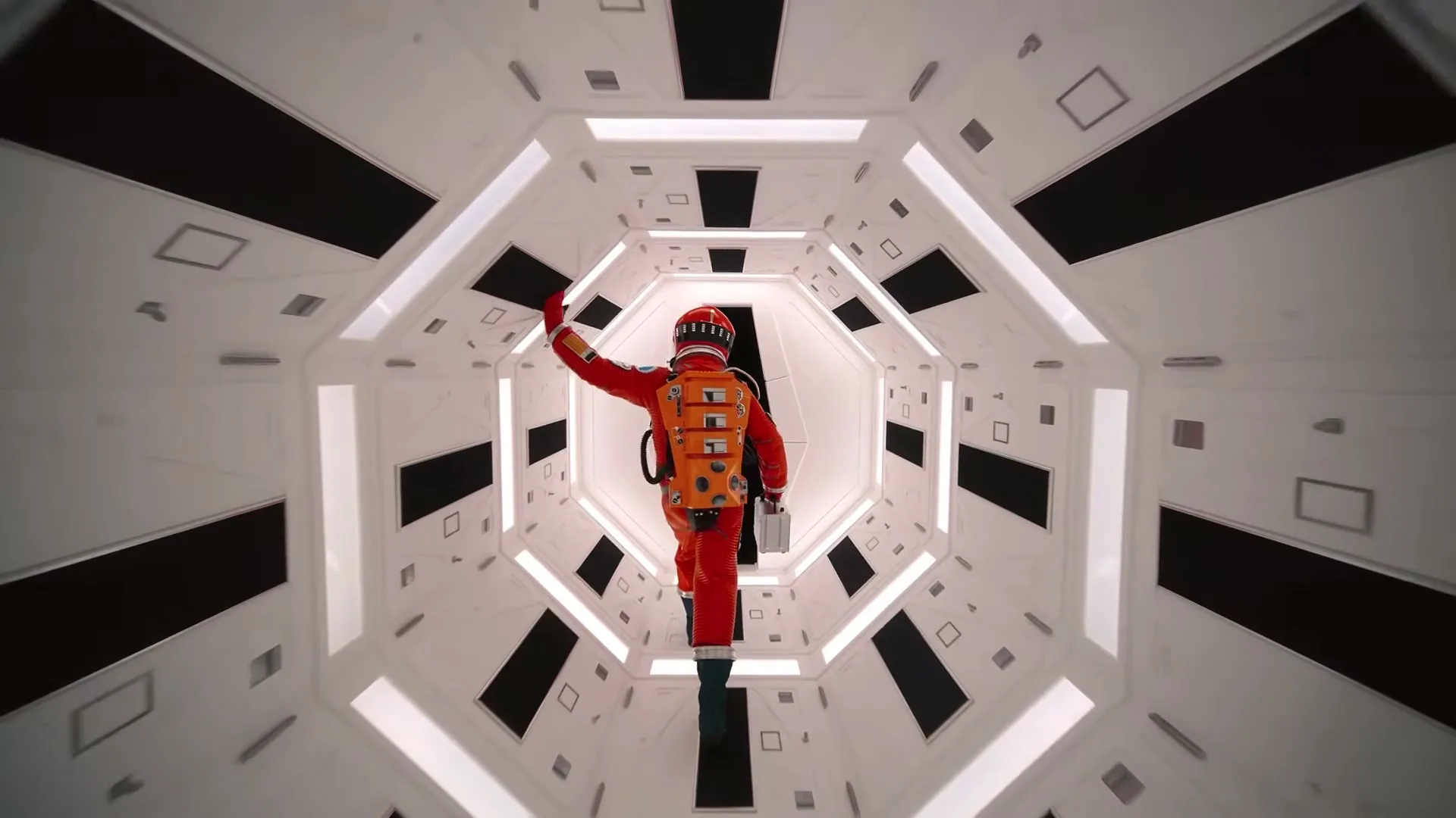 A person in a red spacesuit with an orange backpack floats down a white, geometric spaceship corridor, waving their right hand.