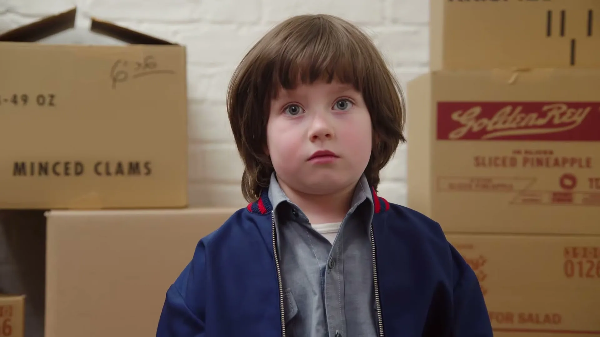 A young boy with a bowl cut and a blue jacket stands in front of stacked cardboard boxes, looking directly at the camera with a neutral expression.