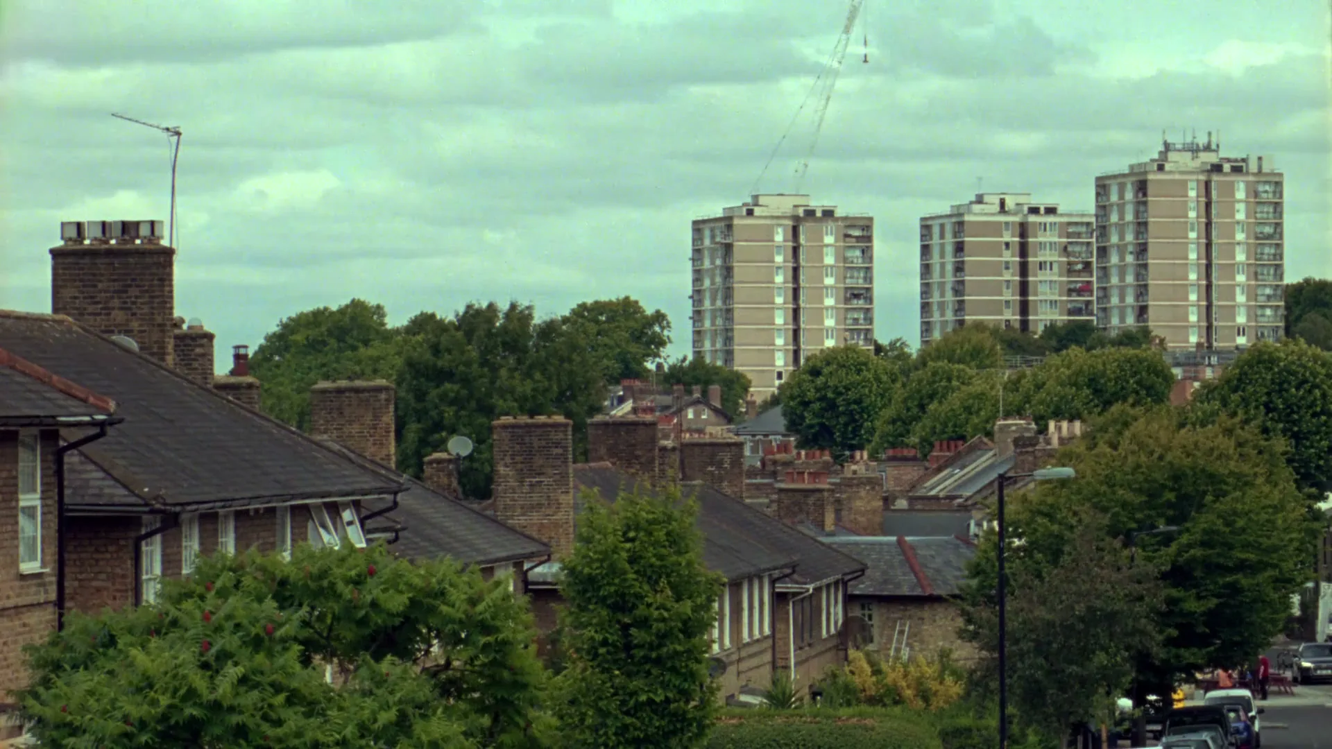 A wide shot of a suburban neighborhood with houses and trees in the foreground, and tall apartment buildings in the background under a cloudy sky.