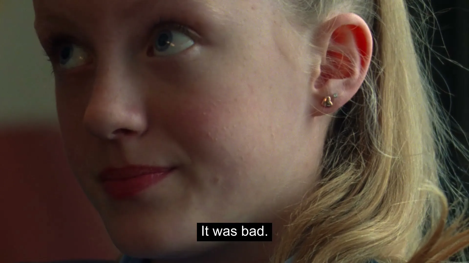 A young blonde girl with red lipstick and a gold earring looks up and then down, with a subtle smile, in an interior setting.