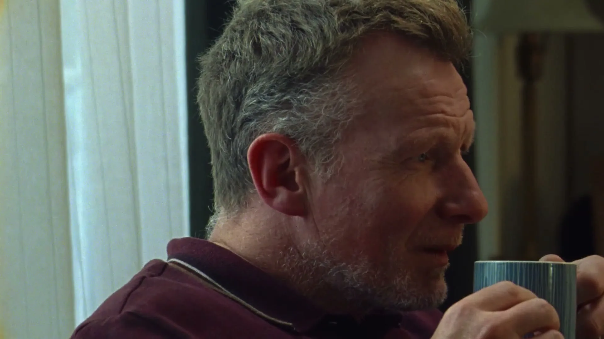 A middle-aged man with reddish-blonde hair and a beard, wearing a maroon polo shirt, holds a blue mug and looks to the side in an interior setting.
