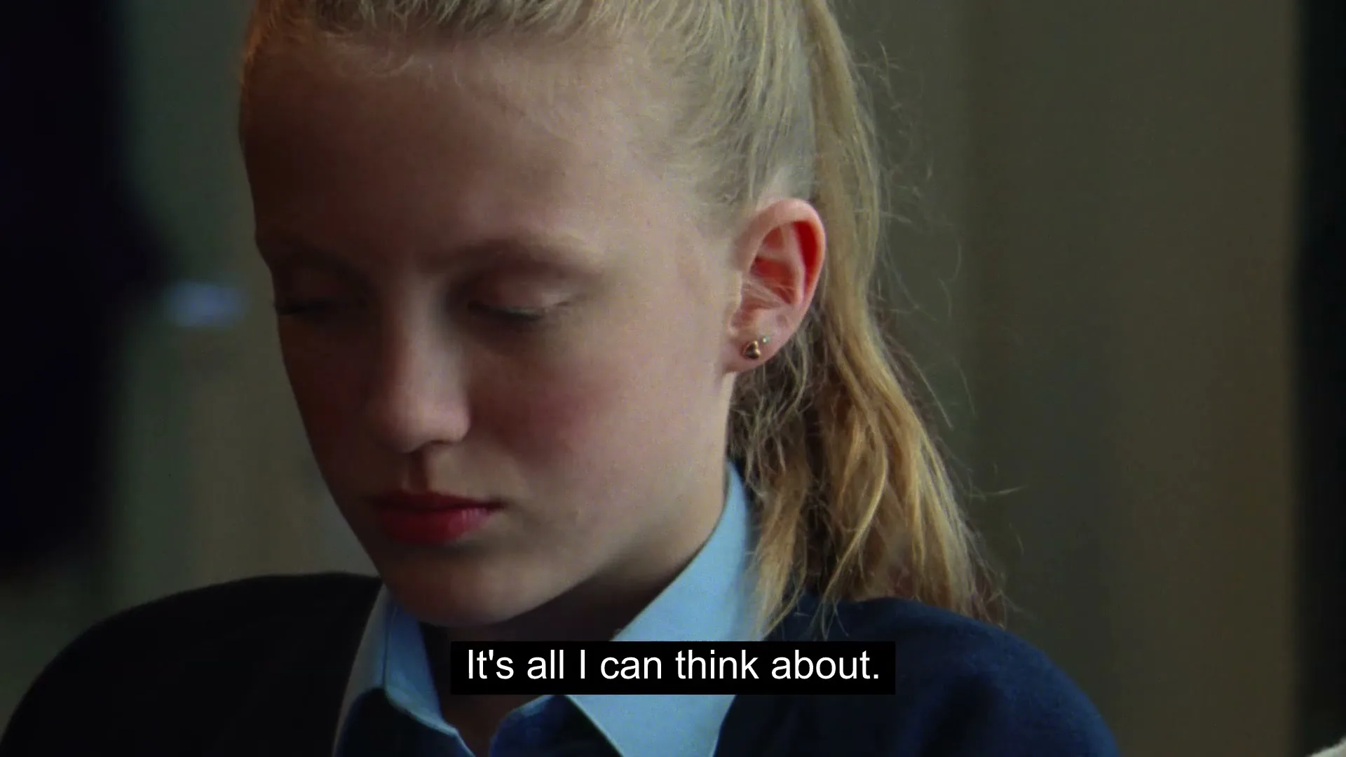 A young blonde girl in a school uniform looks down with a thoughtful expression, as subtitles appear on screen in an interior setting.