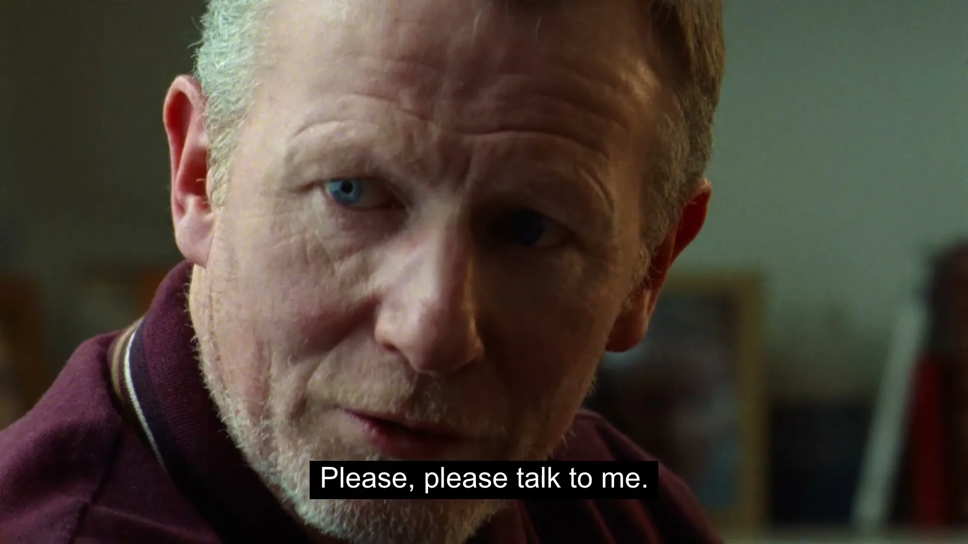 A middle-aged man with blue eyes and a beard looks directly at the camera, wearing a maroon polo shirt in an interior setting.