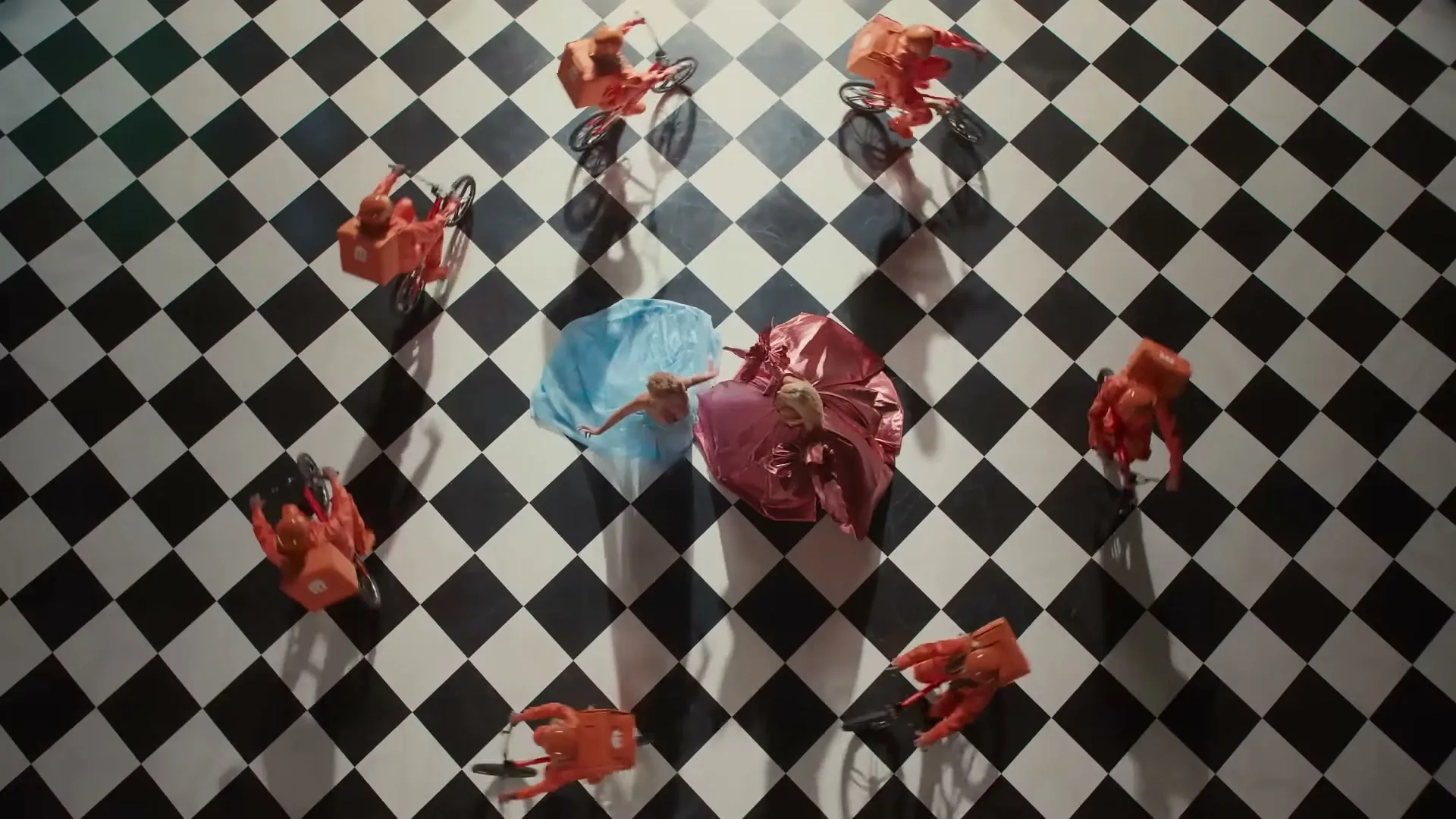 Two women in elaborate dresses dance on a black and white checkered floor, surrounded by several delivery riders on bicycles.