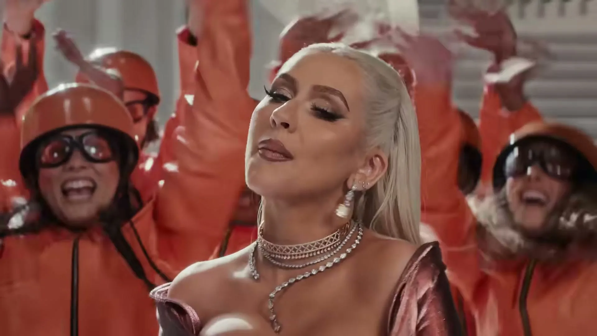 Christina Aguilera, a blonde woman in a sparkly dress and diamond jewelry, sings and dances with a group of people in orange jumpsuits and helmets in a grand hall.