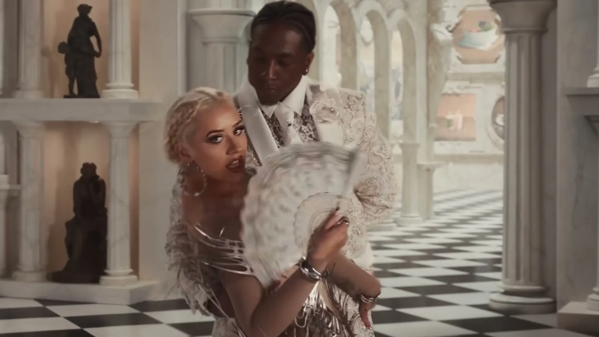 A blonde woman in a silver dress and a Black man in a white suit dance in a grand, ornate hall with a checkered floor.