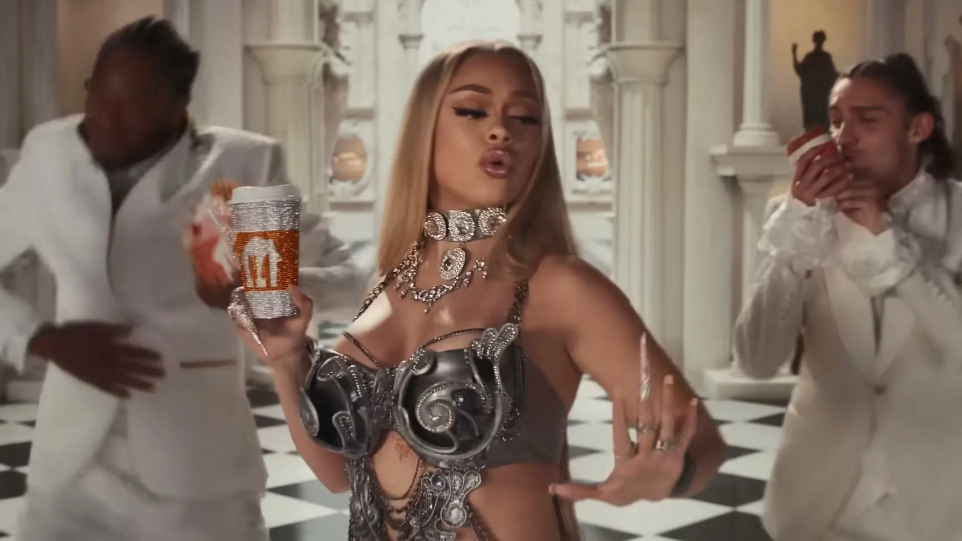 A female rapper in a bejeweled outfit holds a sparkling cup while dancing with two male dancers in a grand hall with a checkered floor.