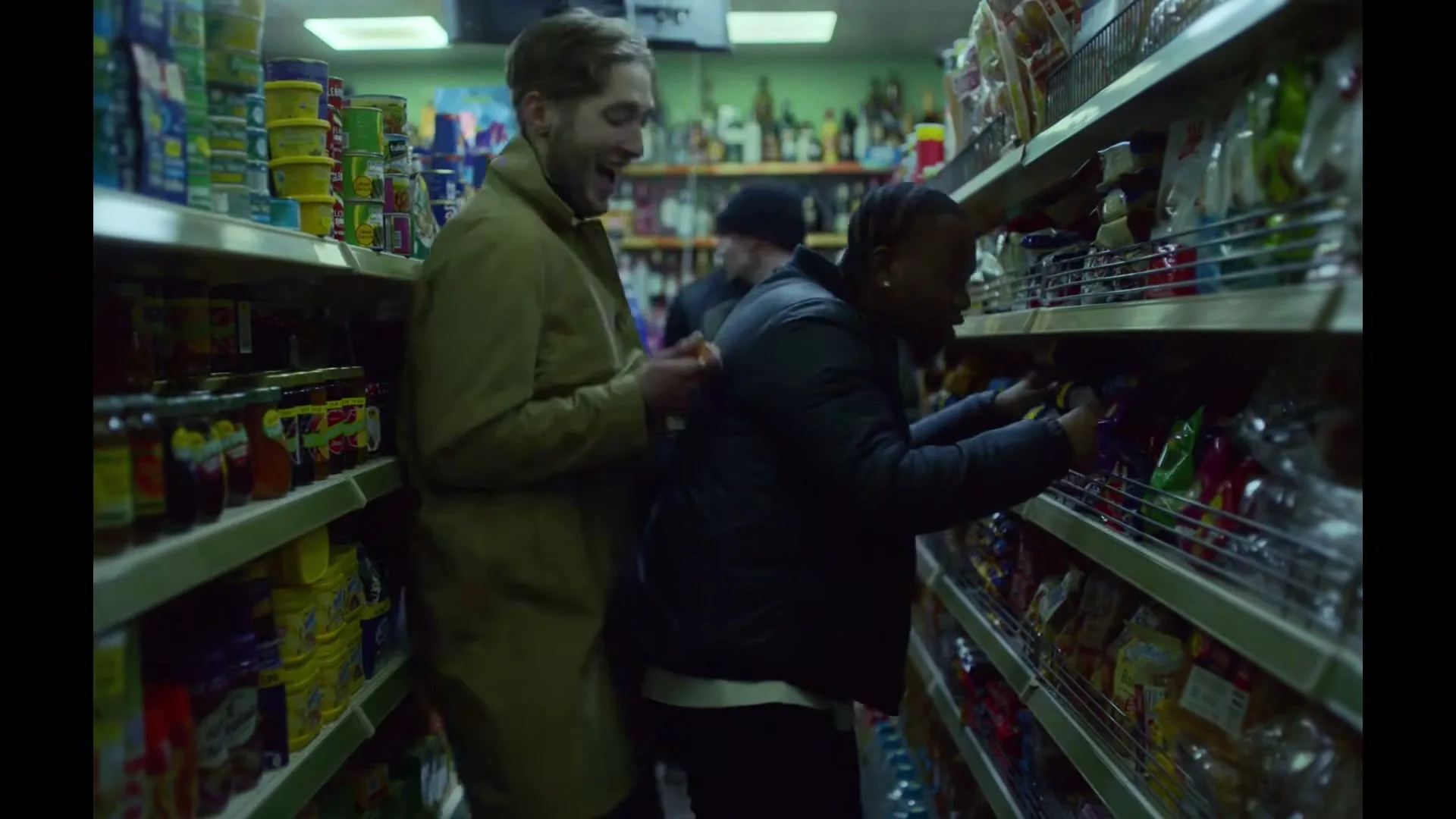 Three young men are in a convenience store aisle, one is shoplifting while another distracts a third, who is laughing.