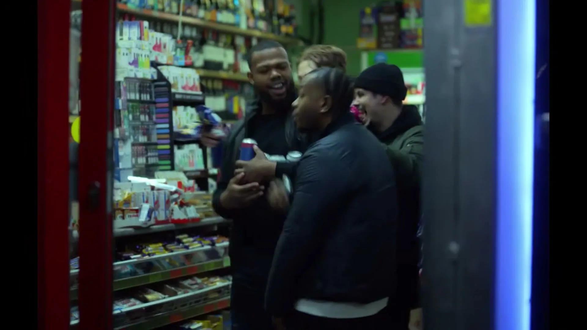 Four young men, two Black and two white, are gathered in a small convenience store, laughing and holding drinks, as one man gestures with a can.