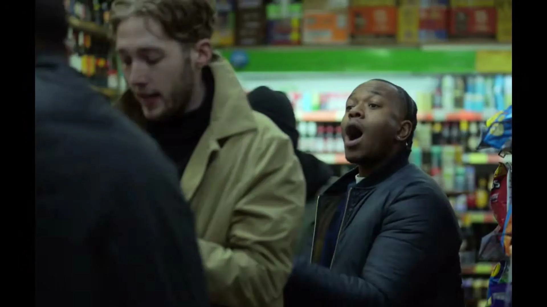 A group of young men are in a convenience store, one man in a tan coat is looking down, while another man in a dark jacket is yelling.