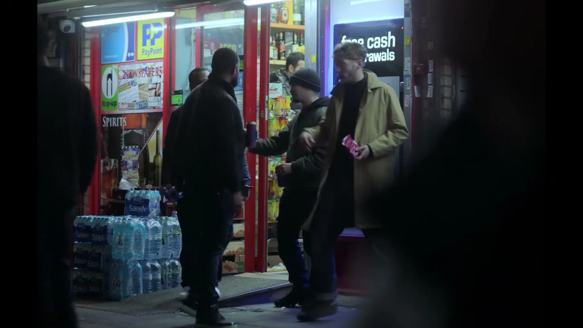 A group of young men stand outside a brightly lit convenience store at night, talking and holding drinks, with one man smoking.