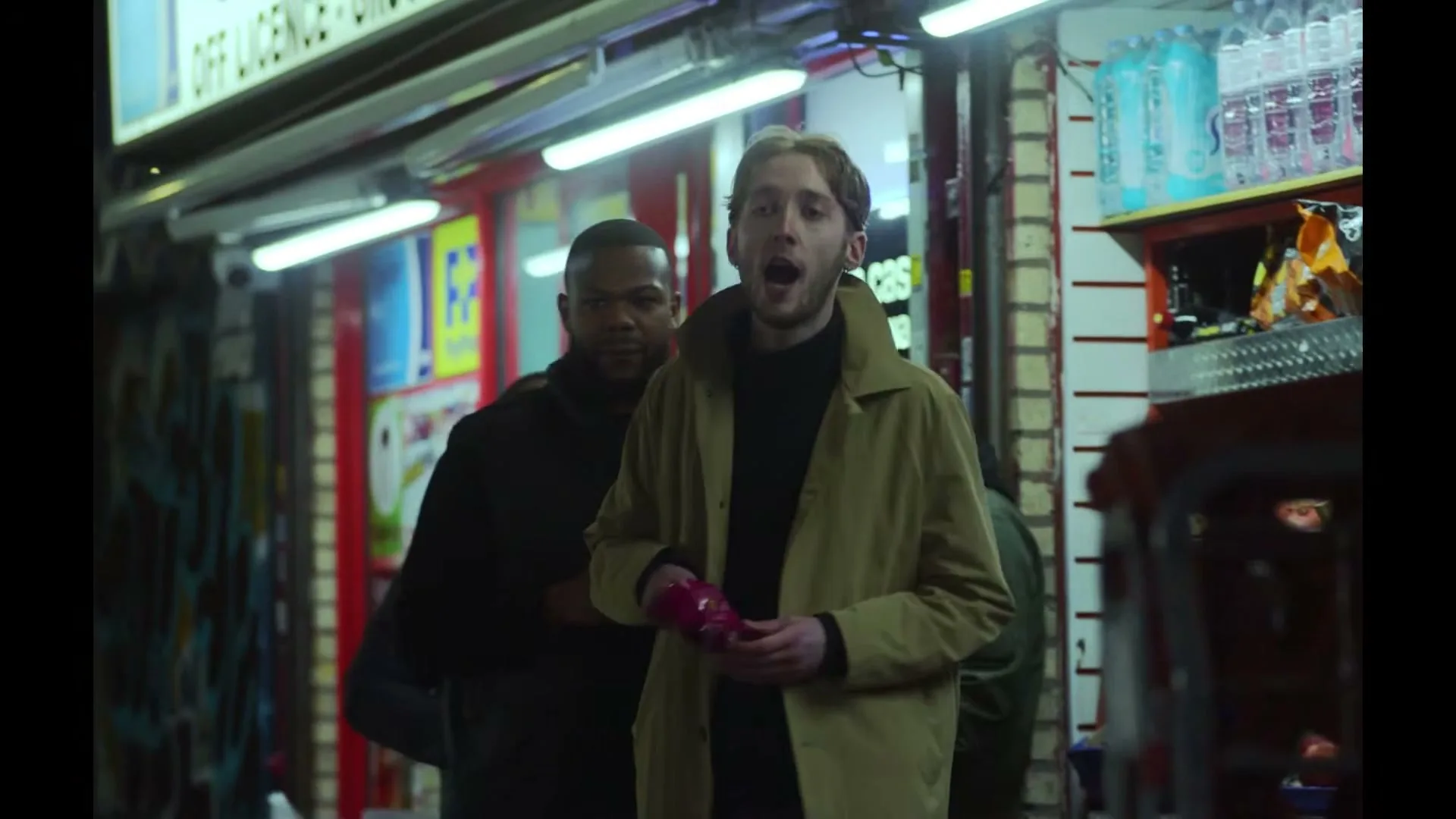 A young man in a trench coat walks down a street at night, holding a pink item, with another man in a black jacket walking behind him.