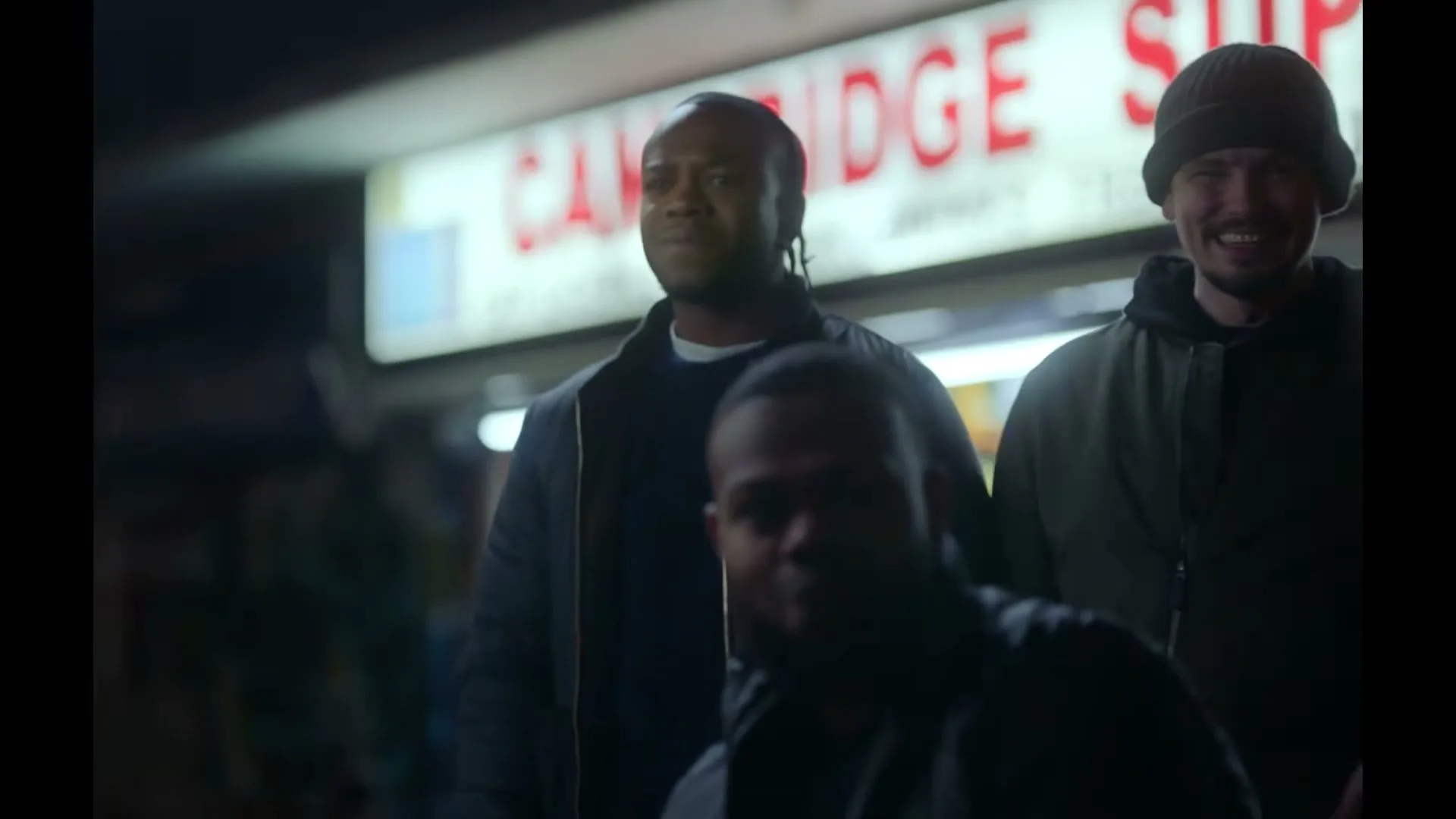 Three men stand outside a shop at night, looking towards the camera with varied expressions, as one man in the foreground smiles.