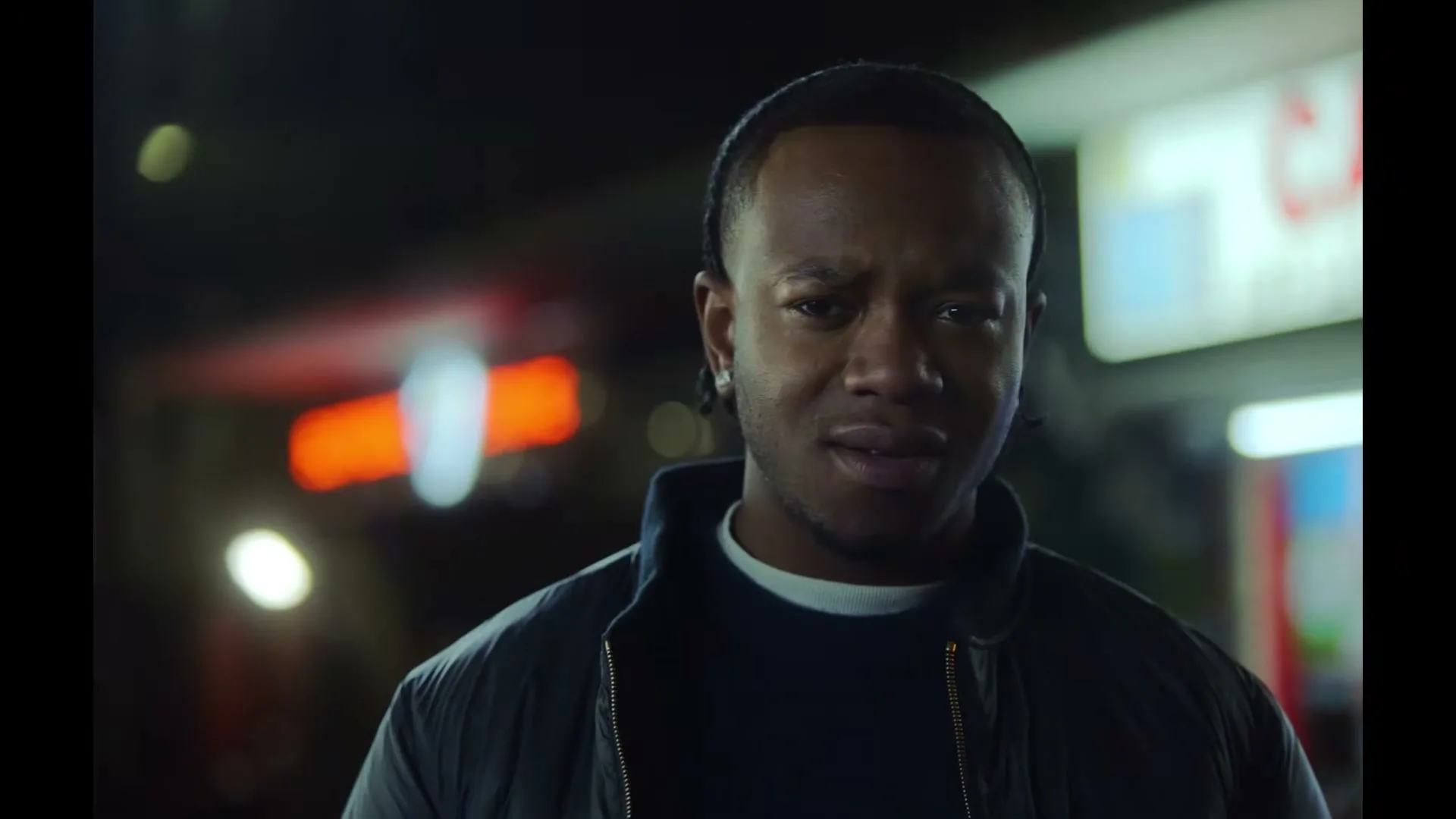 A Black man in his 20s, wearing a dark jacket and sweater, looks directly at the camera with a disgusted expression on a city street at night.