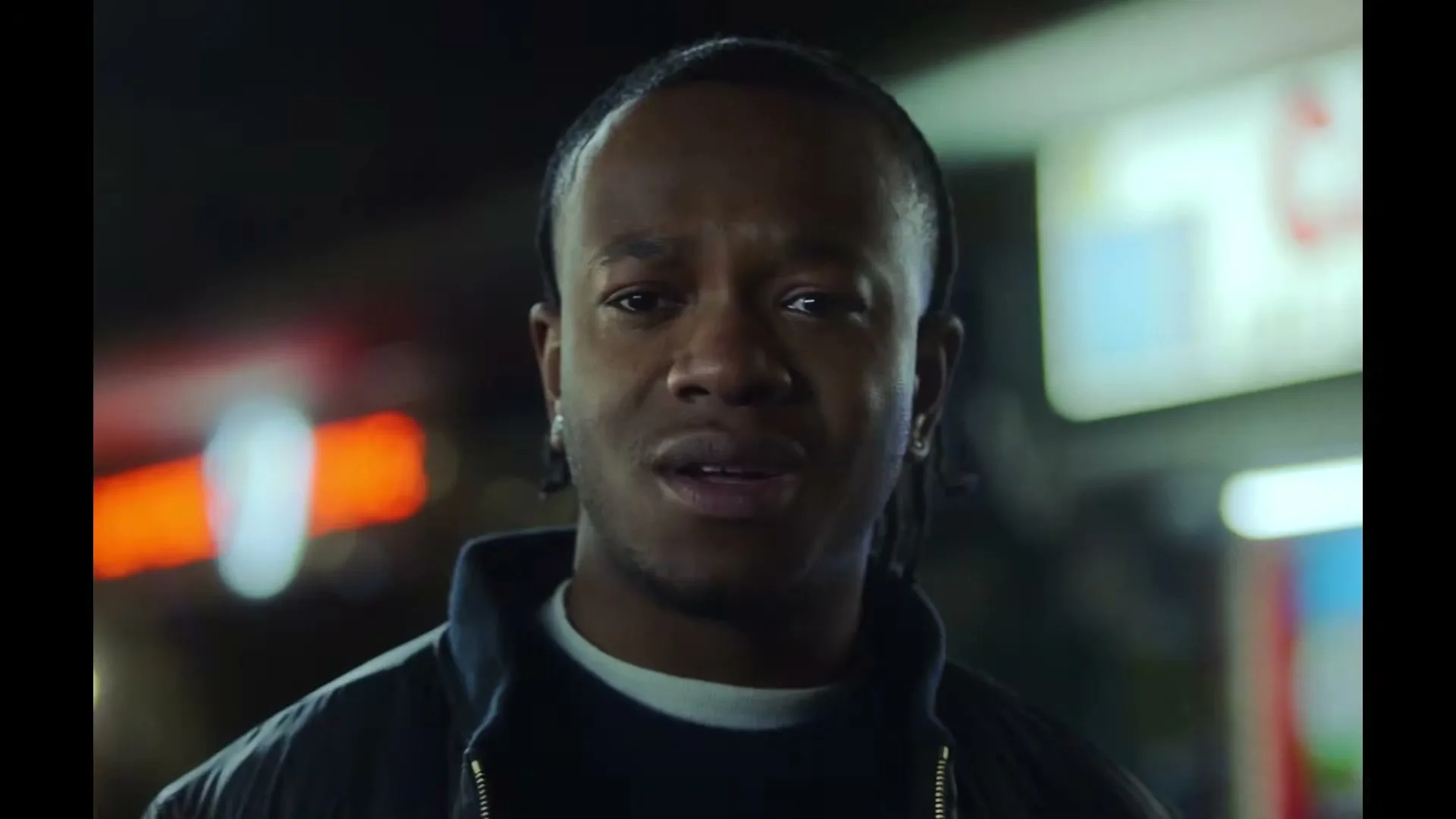 A young Black man with braided hair looks directly at the camera with a concerned expression on a city street at night.