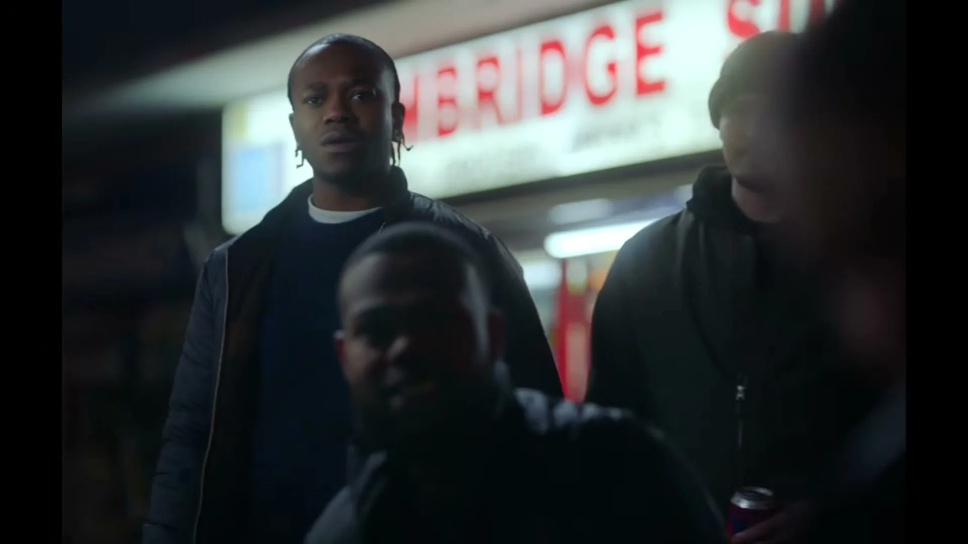 Three men stand outside a shop at night, with one man in the foreground out of focus, while the other two look on with serious expressions.