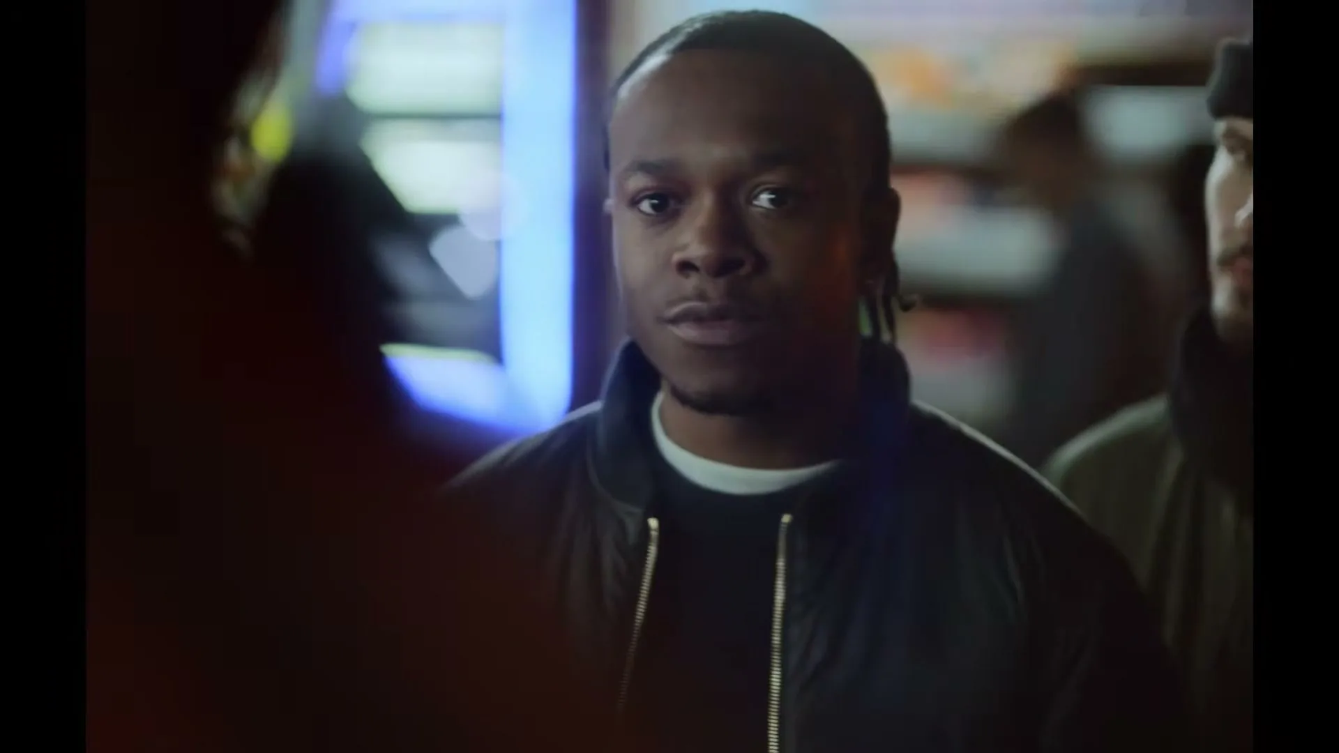 A young Black man with dreadlocks and a dark jacket looks intently at someone off-screen in a dimly lit convenience store.