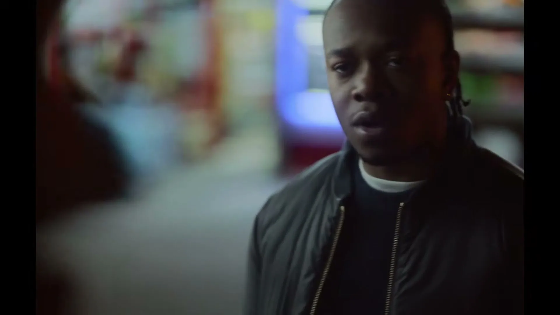 A young Black man with braids, wearing a dark jacket, looks concerned as he speaks to someone off-screen in a dimly lit interior.