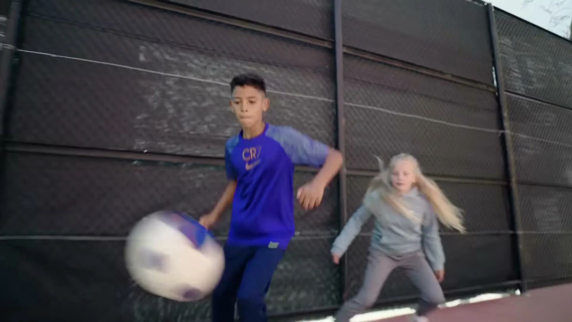 A young boy in a blue shirt dribbles a soccer ball while a young girl runs beside him on a tennis court.