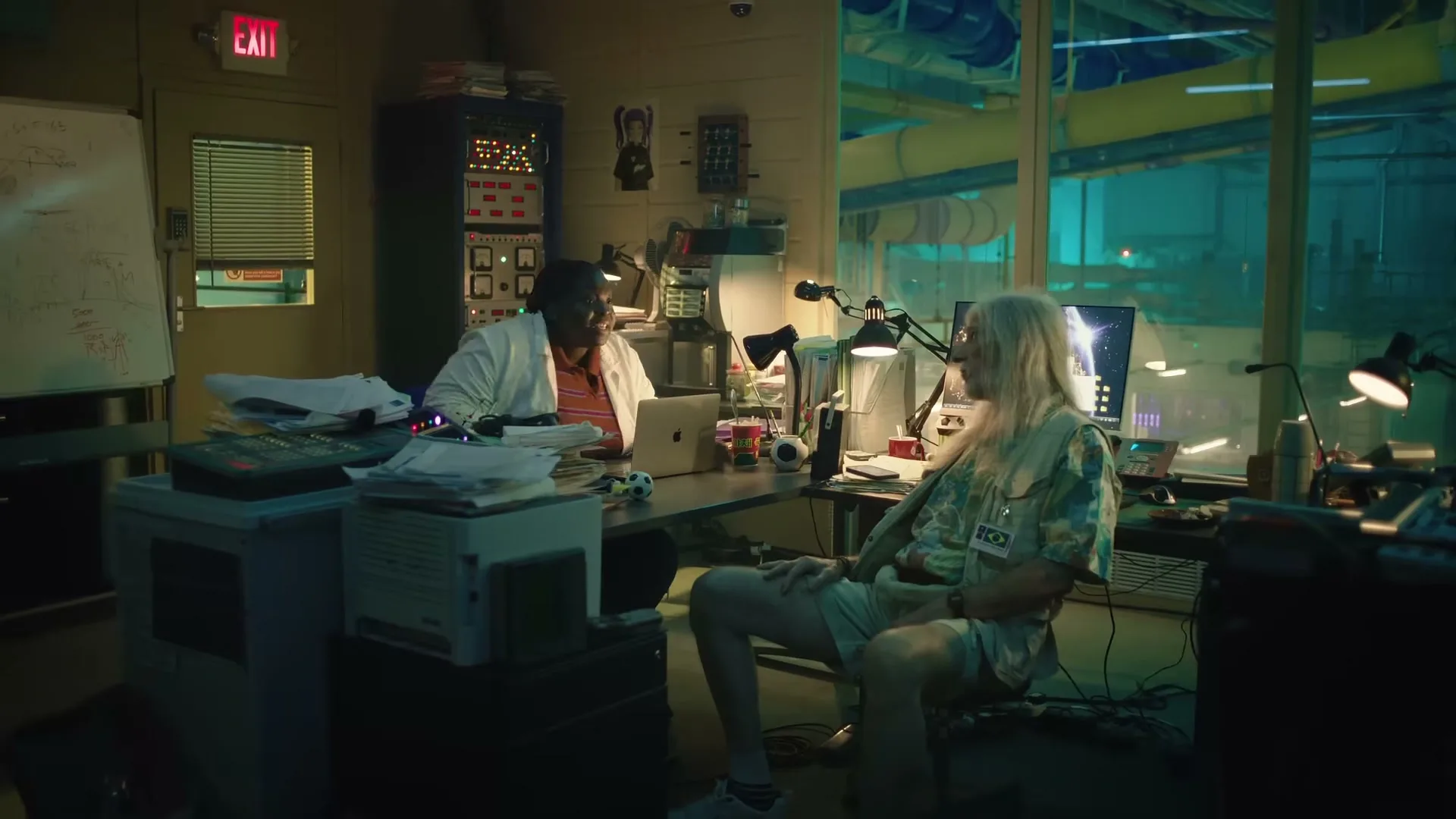 Two men, one in a lab coat and one with long blonde hair, sit at a cluttered desk in a dimly lit office with a water park visible outside.