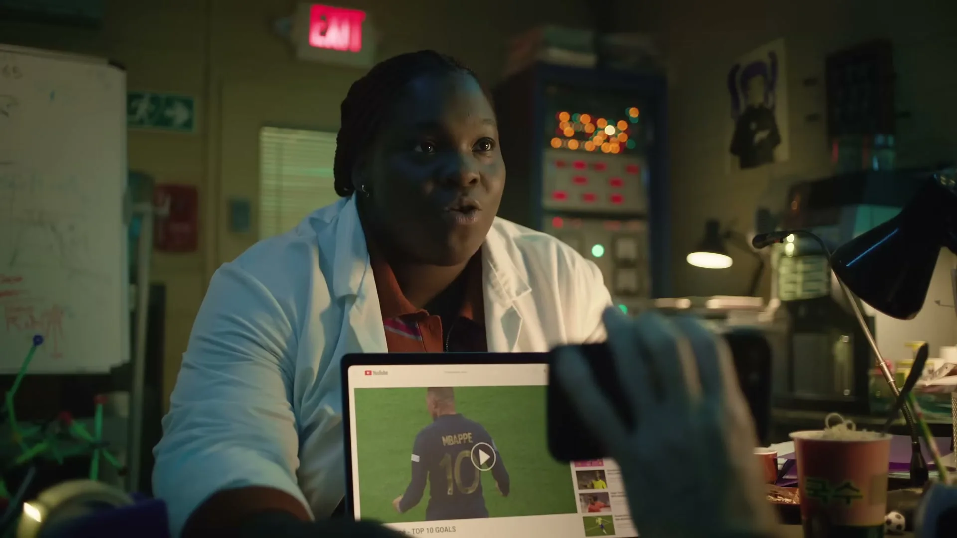 A female scientist in a lab coat talks to someone off-screen, while a video of Kylian Mbappé plays on a laptop in a cluttered lab.