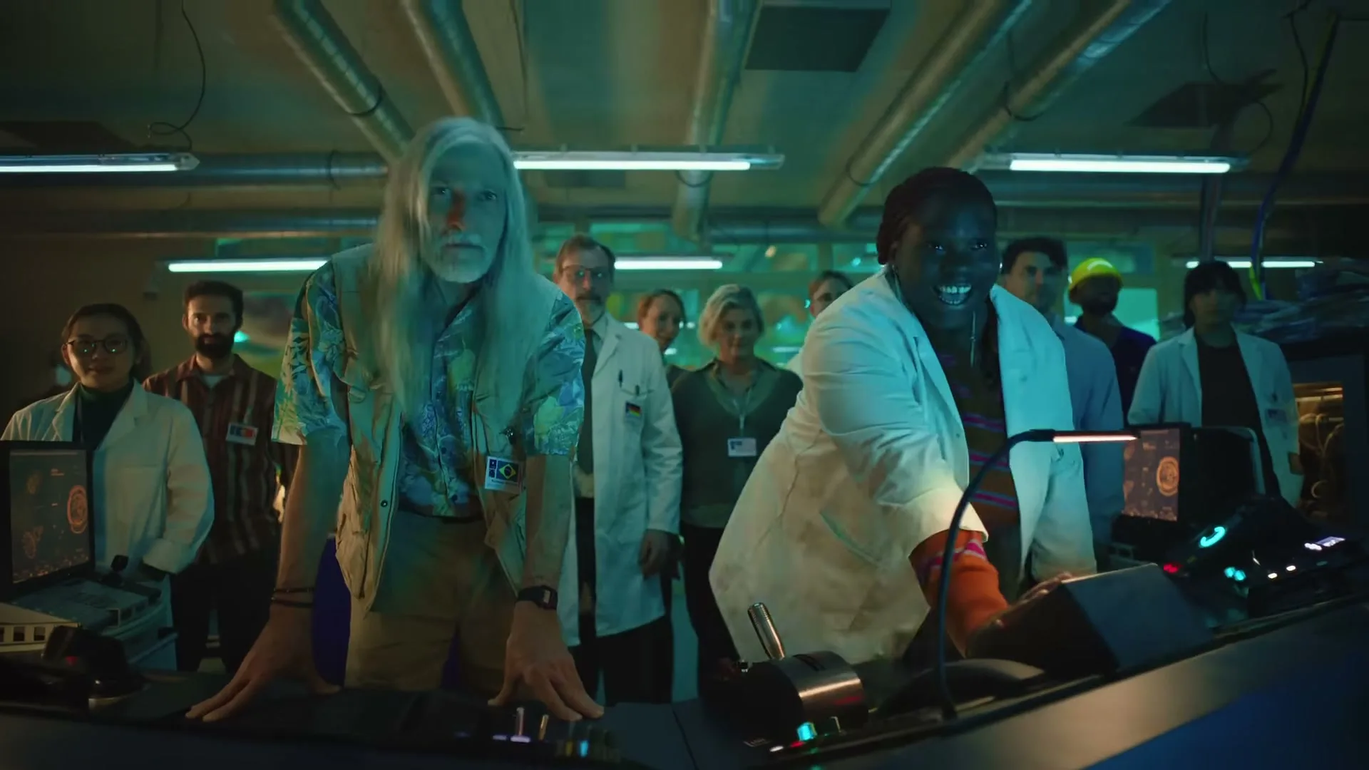 A diverse group of scientists and technicians, including an older man with long white hair and a younger Black woman, stand in a control room, looking surprised and excited.