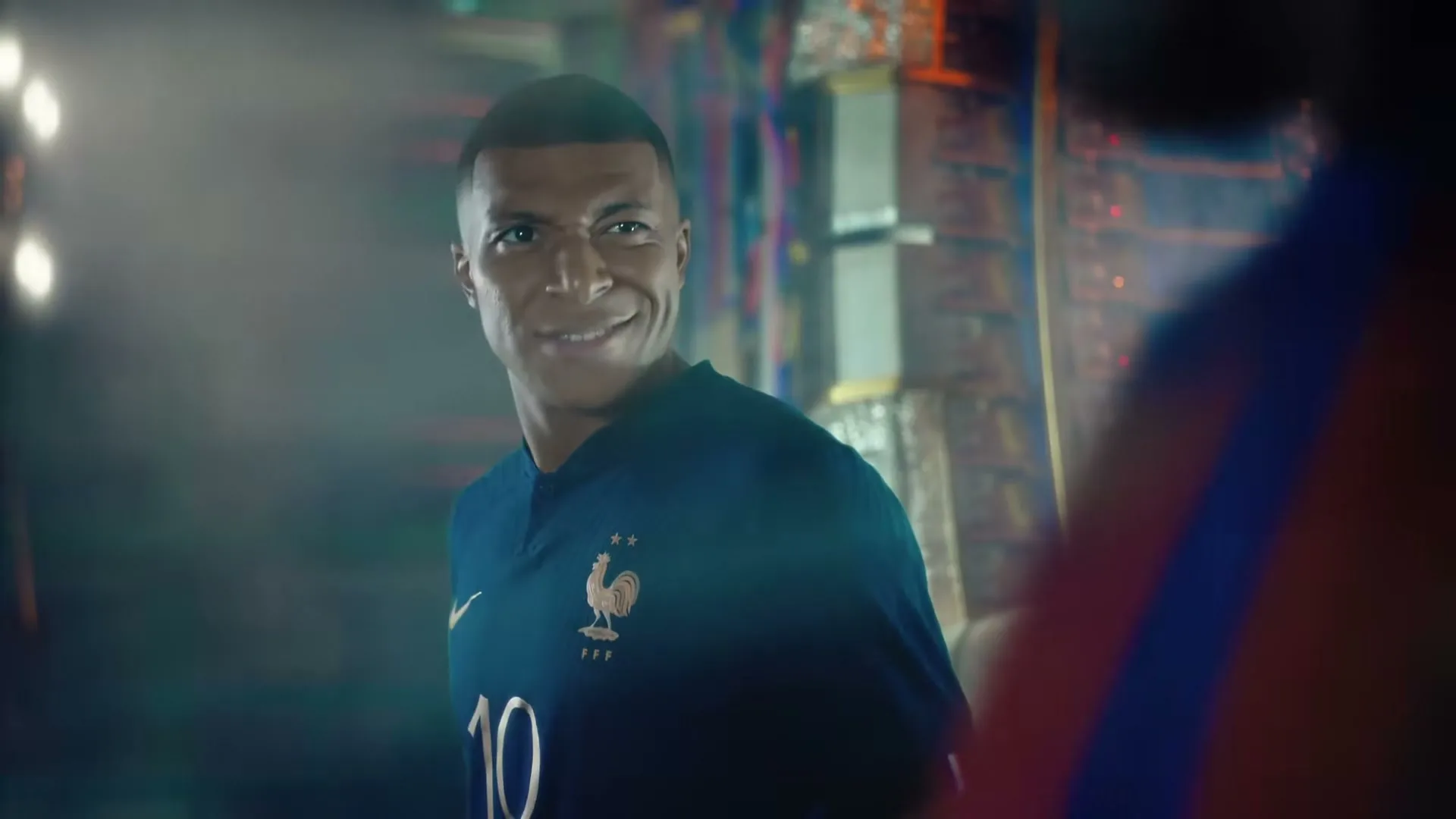 Kylian Mbappé, wearing a blue France national football team jersey, looks at someone off-screen and smiles in a locker room.