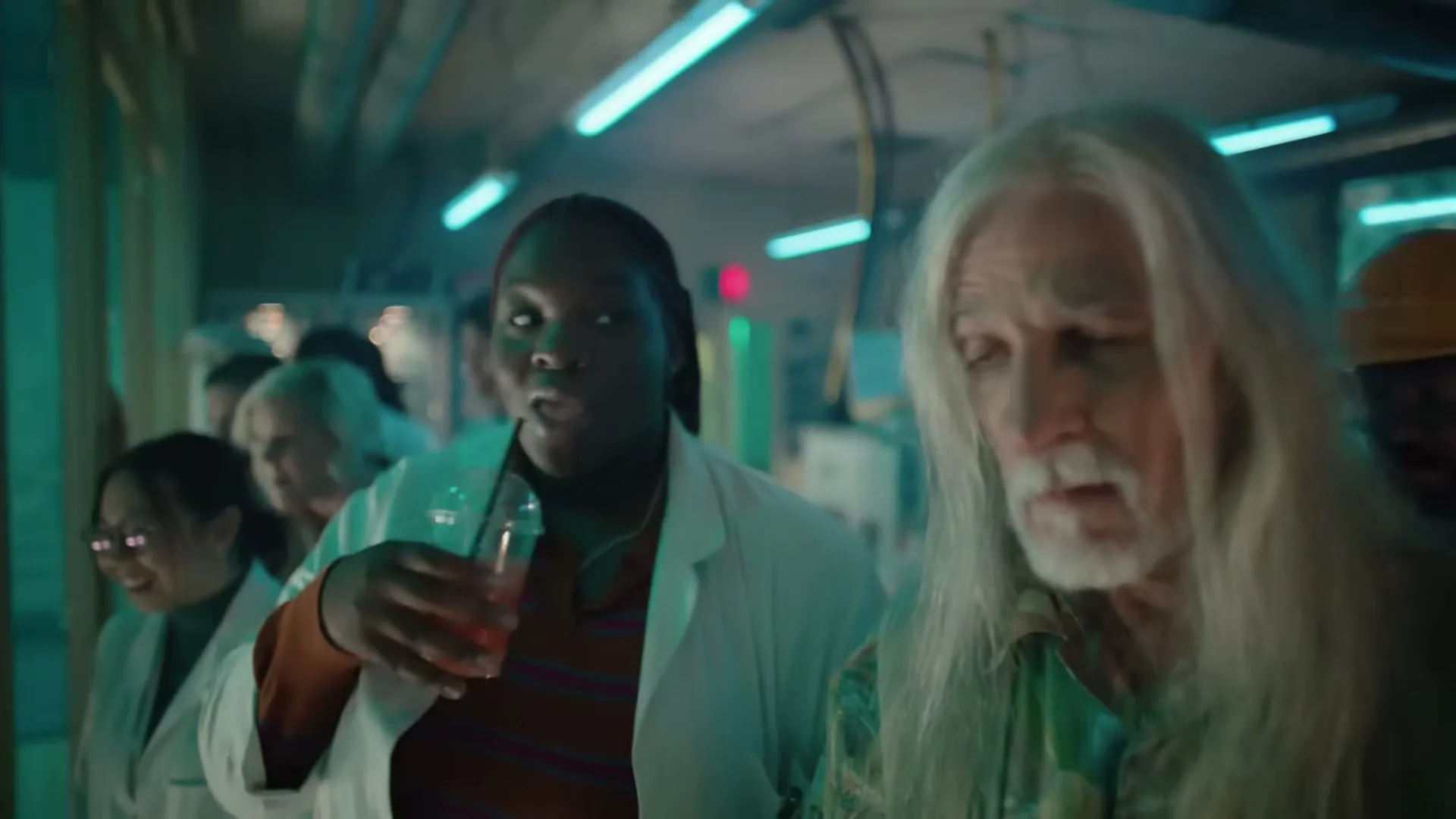 A diverse group of people, including a woman in a lab coat and an older man with long white hair, watch a soccer game in a dimly lit, futuristic lab.